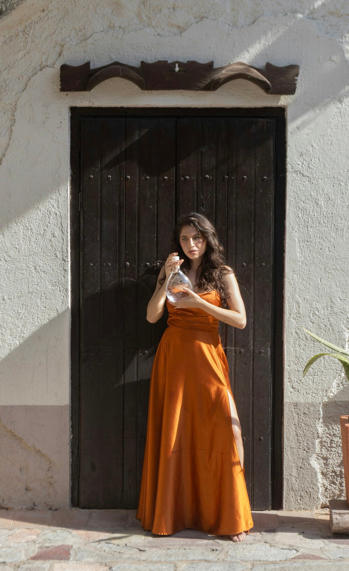 Mujer con vestido largo de satín naranja frente a un portón de madera oscura, en un muro encalado.