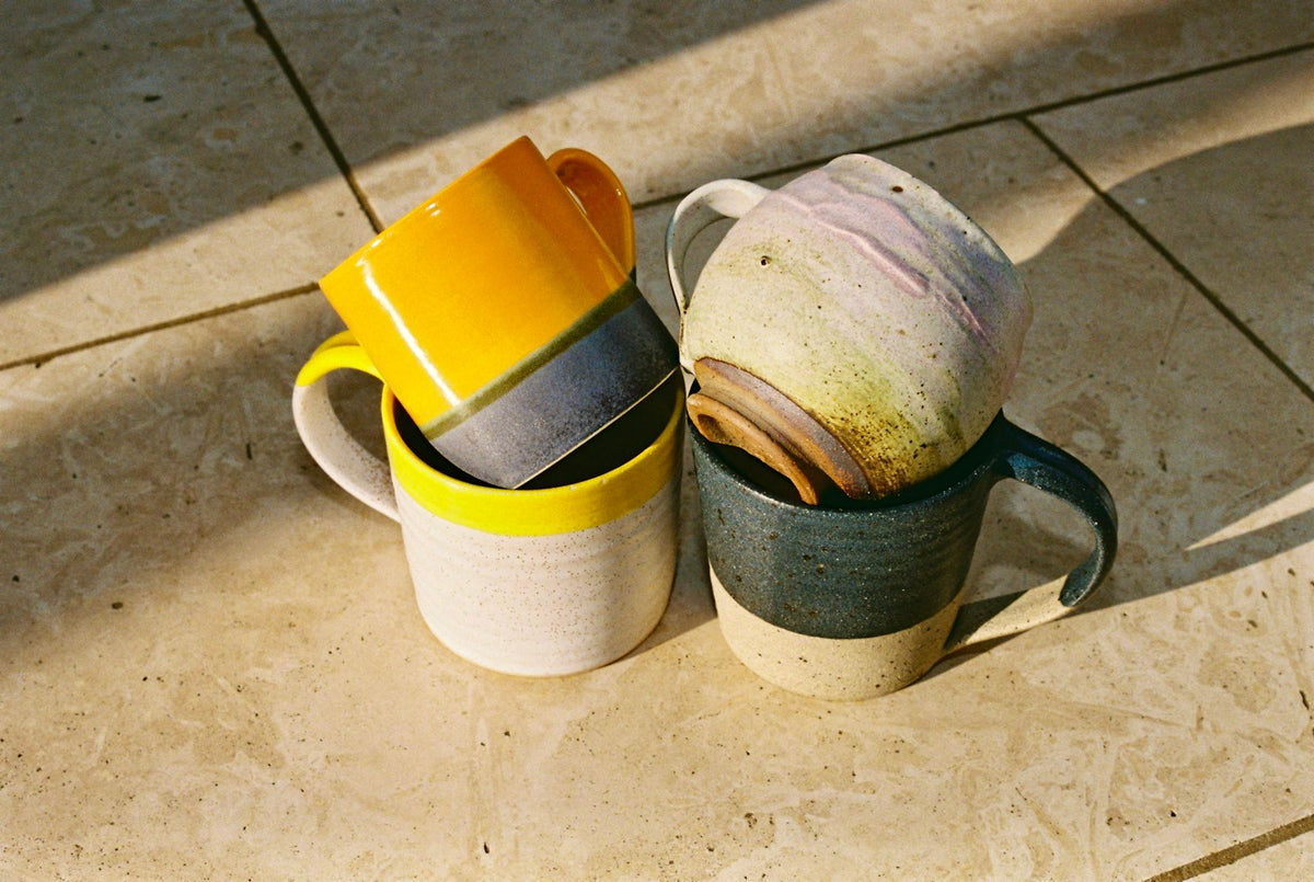 Tazas de cerámica esmaltadas en amarillo, gris y verde, juntas sobre un piso de piedra a pleno sol.