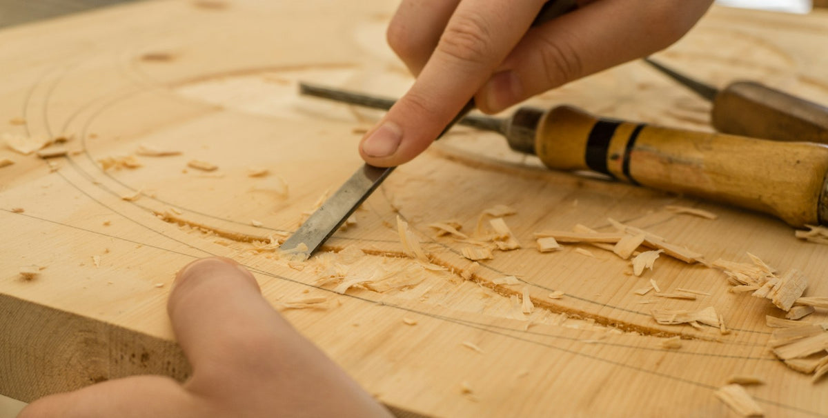 Unas manos tallan una tabla de madera clara con una gubia; hay virutas sobre la mesa.