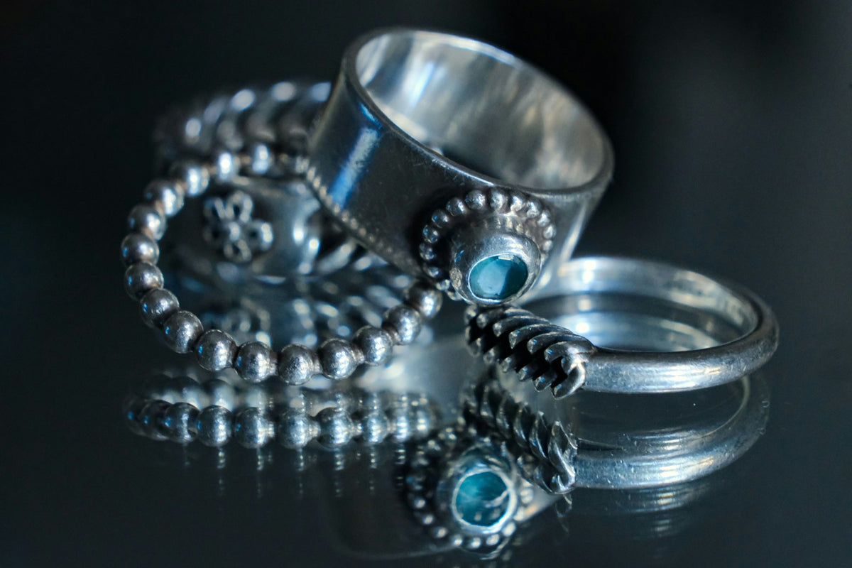 Anillos de plata trabajada, dos con turquesas, sobre una superficie oscura que los refleja.