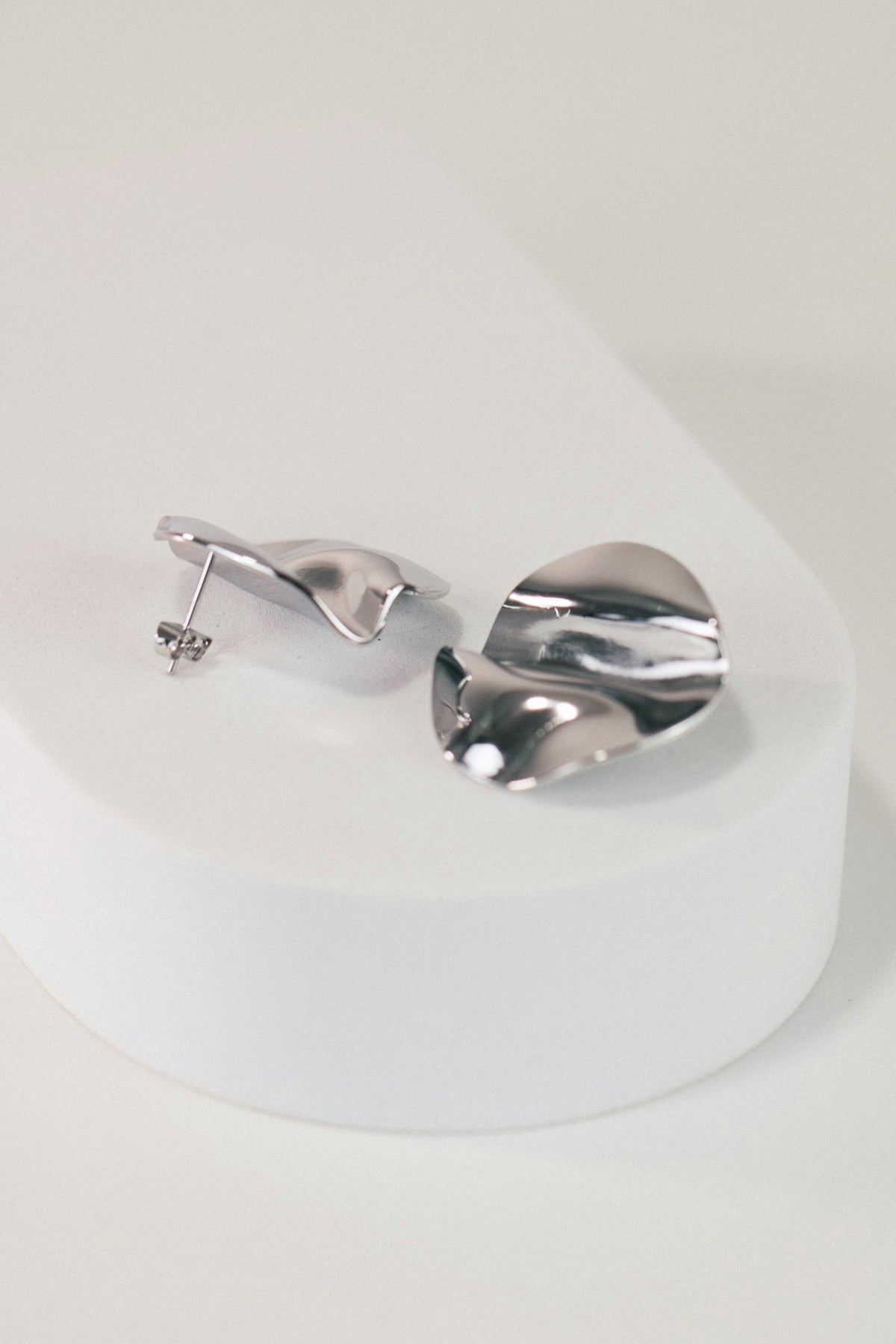Par de aretes de plata ondulada como pétalos, sobre una base blanca.