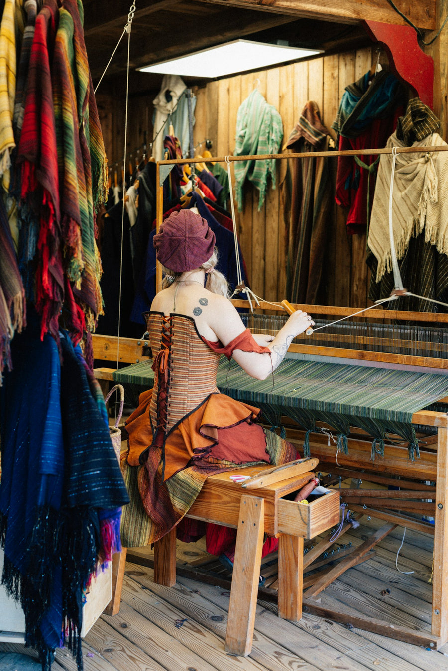 Una tejedora trabaja en un telar de pedal, rodeada de textiles de colores.