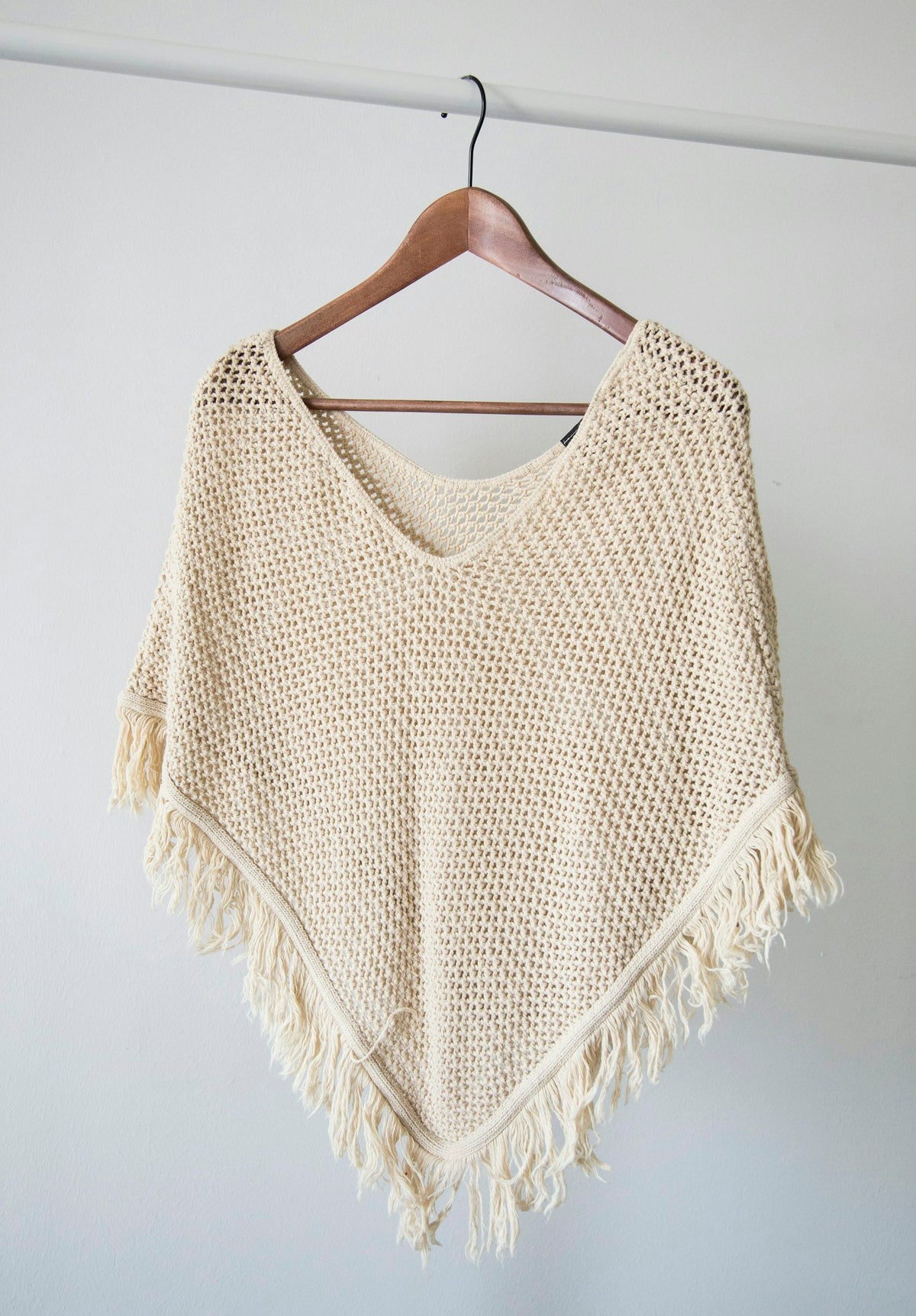 Poncho tejido de algodón color crudo, con flecos, colgado en un gancho de madera.