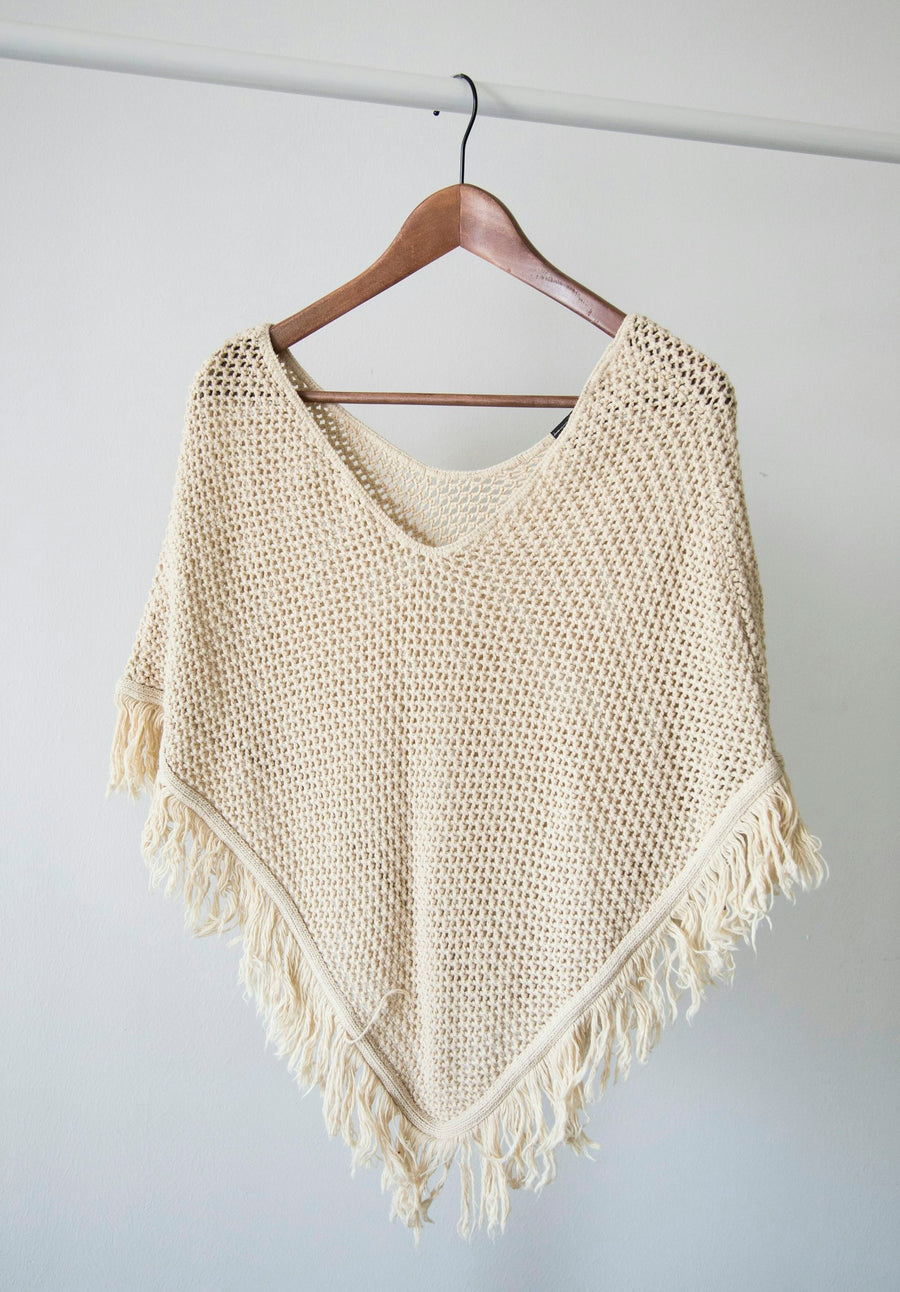 Poncho tejido de algodón color crudo, con flecos, colgado en un gancho de madera.