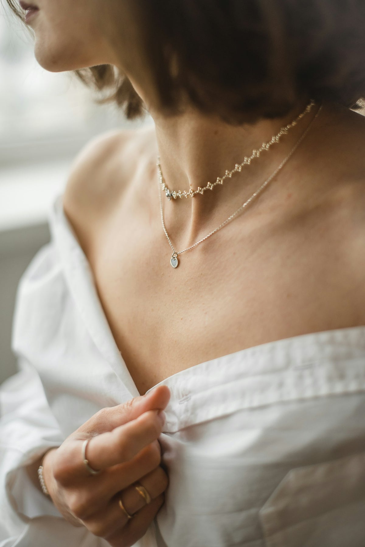 Mujer con camisa blanca abierta y dos collares finos de plata, uno de cuentas claras y otro con una gota.