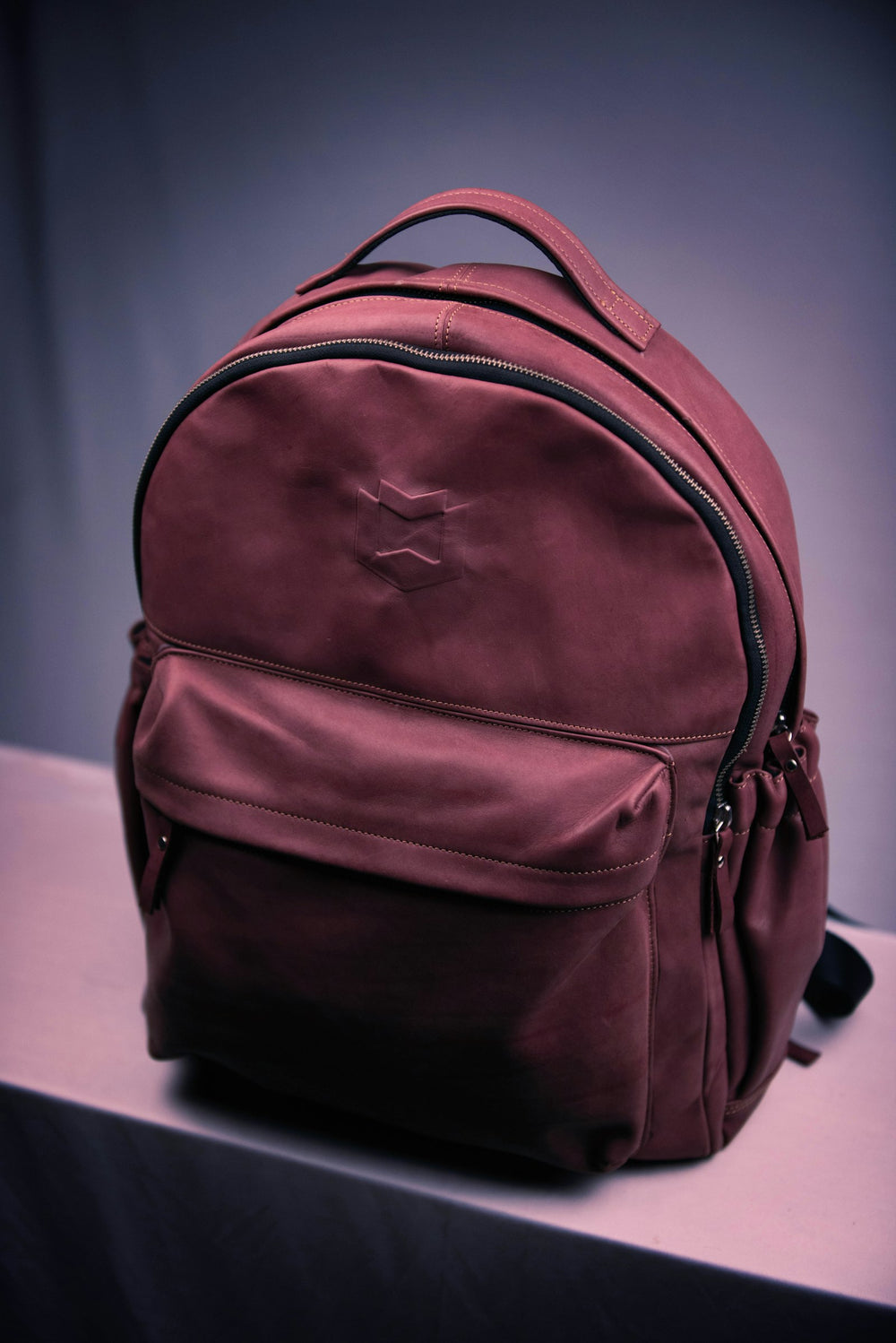 Mochila de piel color vino con una bolsa al frente, sobre una mesa clara y contra un fondo lila.