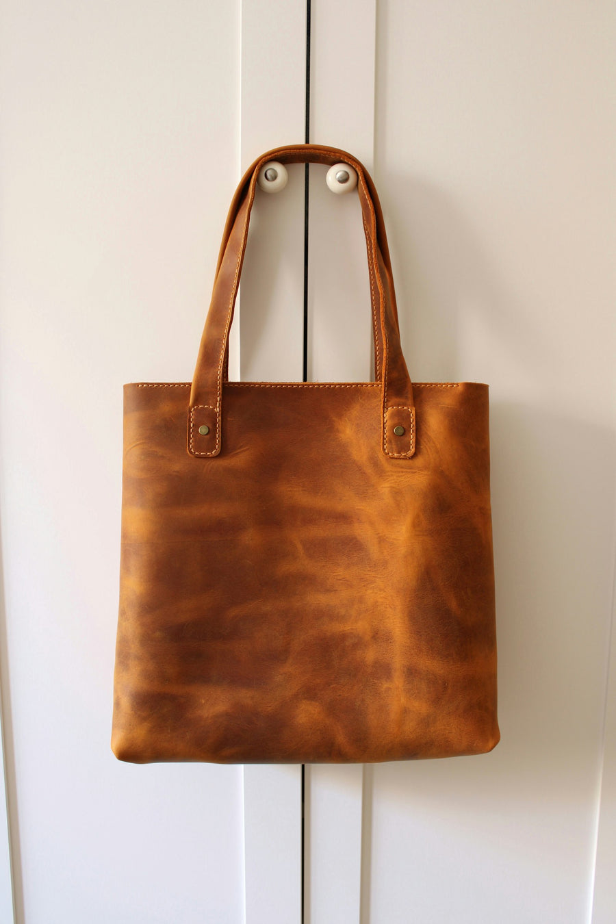 Bolsa tote de piel color miel, de costuras visibles, colgada en un muro blanco.