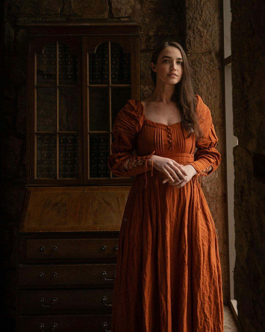 Mujer con vestido largo de lino color terracota y mangas abullonadas, en un cuarto en penumbra.
