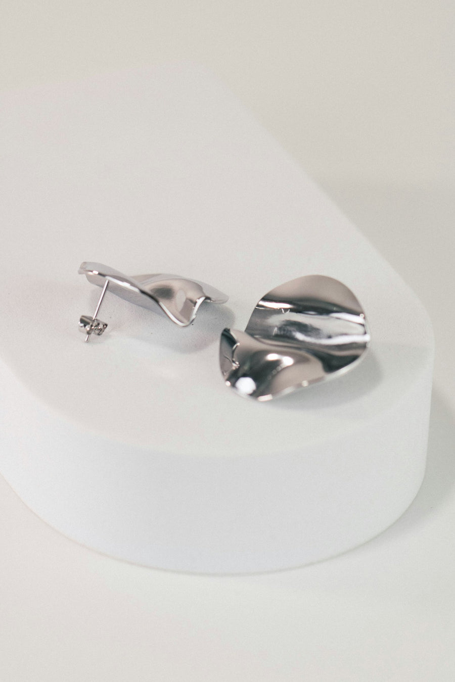 Par de aretes de plata ondulada como pétalos, sobre una base blanca.