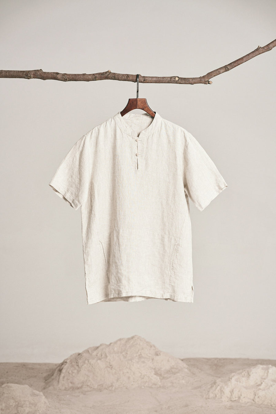 Camisa de manta color crudo, colgada de una rama sobre arena clara.