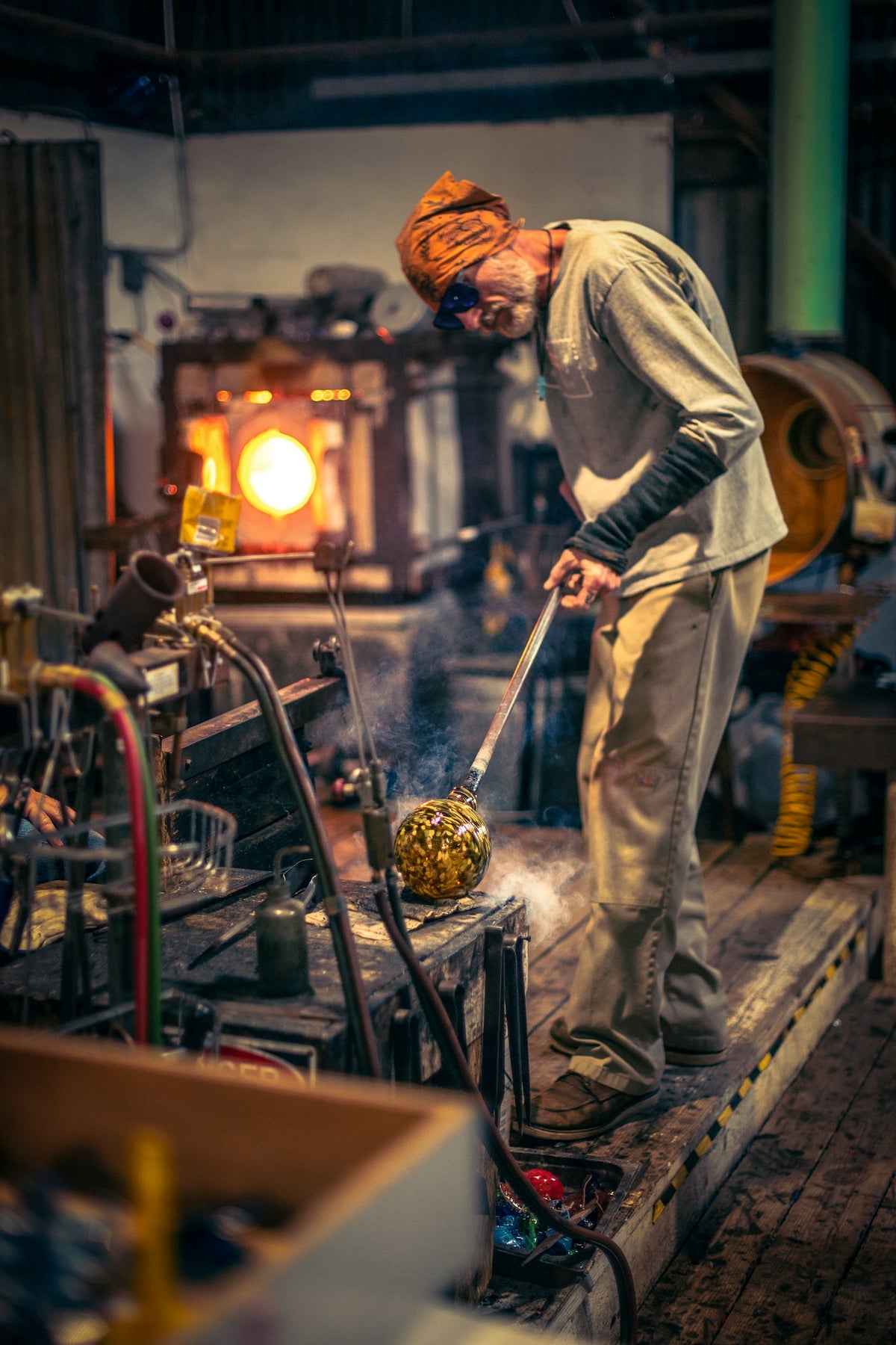 Un vidriero con paliacate naranja trabaja una bola de vidrio al rojo frente al horno de su taller.