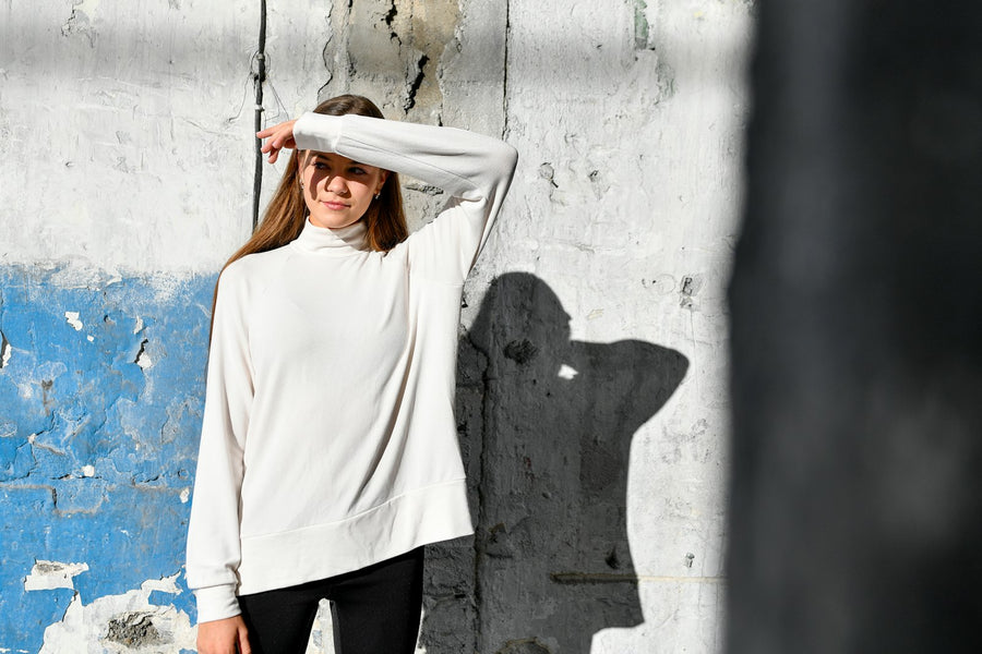 Mujer con suéter blanco de algodón frente a un muro encalado cruzado por sombras.